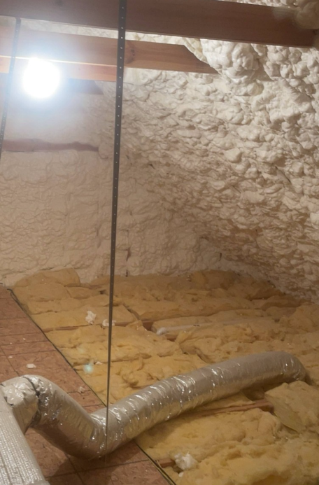 Attic space with spray foam insulation on the roof and walls, yellow batt insulation on the floor, and flexible HVAC ductwork running across the area.