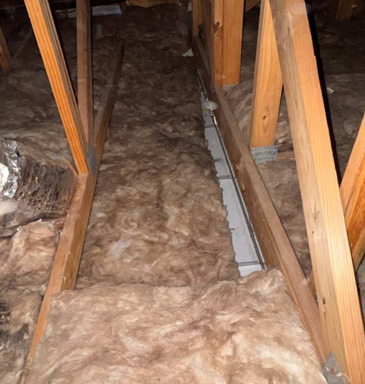 Attic area with wooden truss framing and batt insulation laid between the joists, with HVAC ductwork partially visible on one side.
