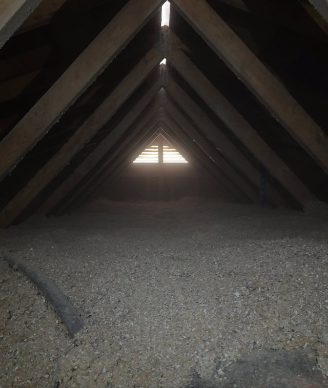 Attic space with triangular roof framing and blown-in insulation covering the floor, illuminated by light coming through a vent at the far end.