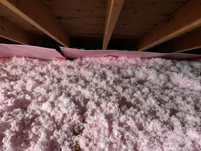 Thick pink blown-in fiberglass insulation covering an attic floor, topped with pink attic insulation baffles positioned beneath exposed wooden roof rafters.