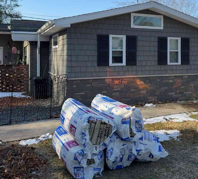 Stacked rolls of packaged R-13 fiberglass insulation sitting on the lawn in front of a grey, residential home with dark shutters during an insulation installation project by Reed's Home Solutions.