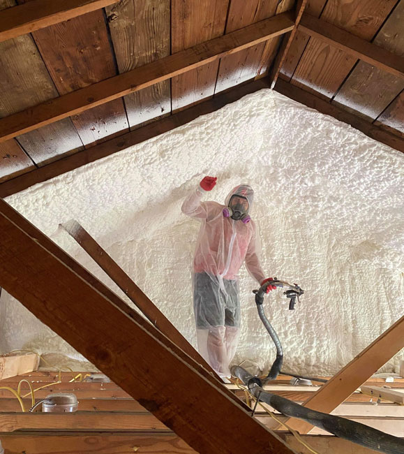 An insulation technician wearing full safety equipment, including a protective suit and respirator mask, posing with a spray foam hose inside an attic featuring newly applied spray foam insulation on the end wall.