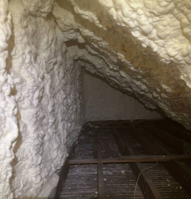 An attic interior showing off-white spray foam insulation freshly applied along the sloped ceiling and walls, covering the wooden frame above dark floor joists.