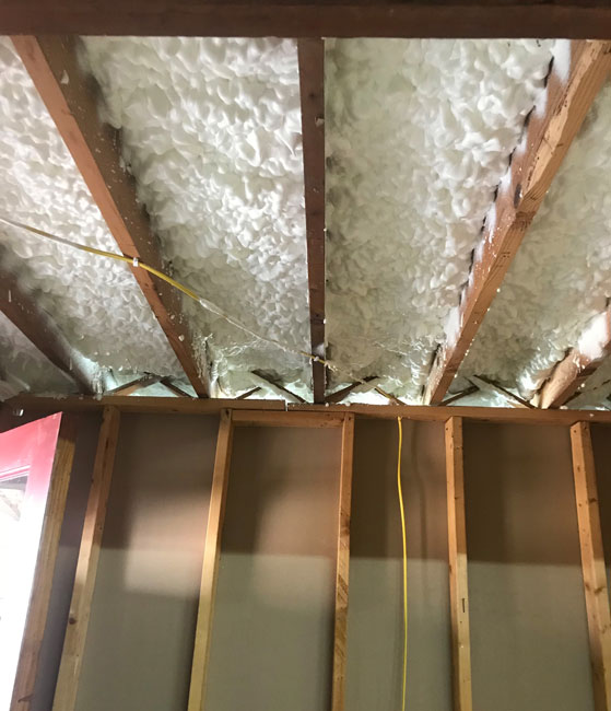 Bright white spray foam insulation applied between exposed wooden ceiling joists above unfinished wall studs in a basement room.
