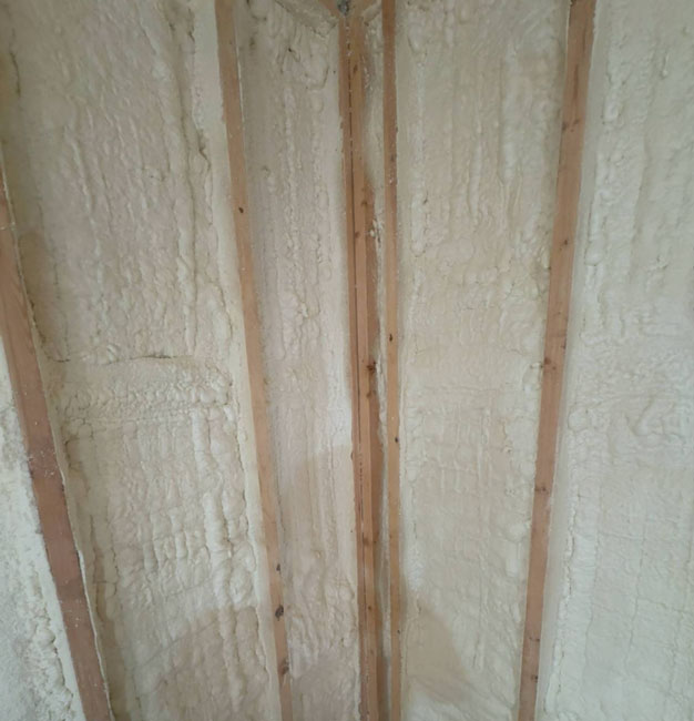 An interior wall corner framed with wooden studs, showing expanding off-white spray foam insulation filling every wall cavity thoroughly.
