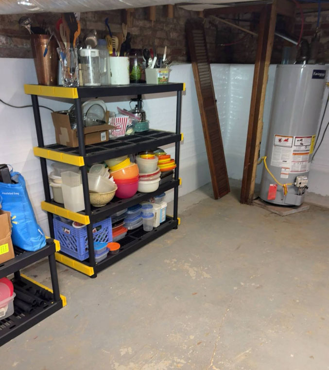 A clean, dry basement space with newly installed white vapor barrier wall panels behind storage shelves loaded with kitchenware and next to a water heater.