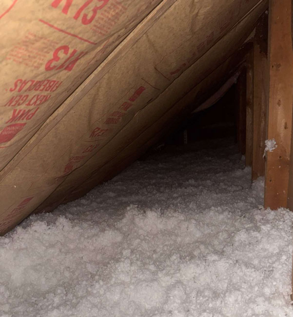 Thick white blown-in fiberglass insulation covering an attic floor space beneath sloped rafters insulated with kraft-faced paper batts.