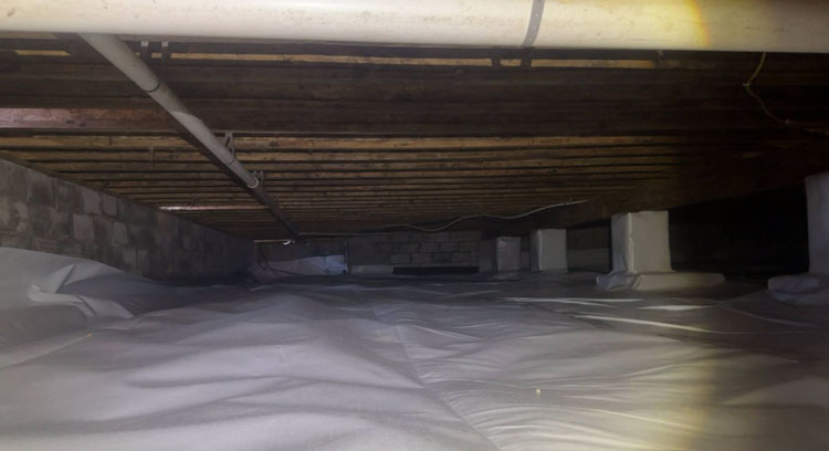 A clean crawl space fully lined with a heavy-duty white vapor barrier across the dirt floor and wrapped around concrete support piers beneath exposed subfloor joists.