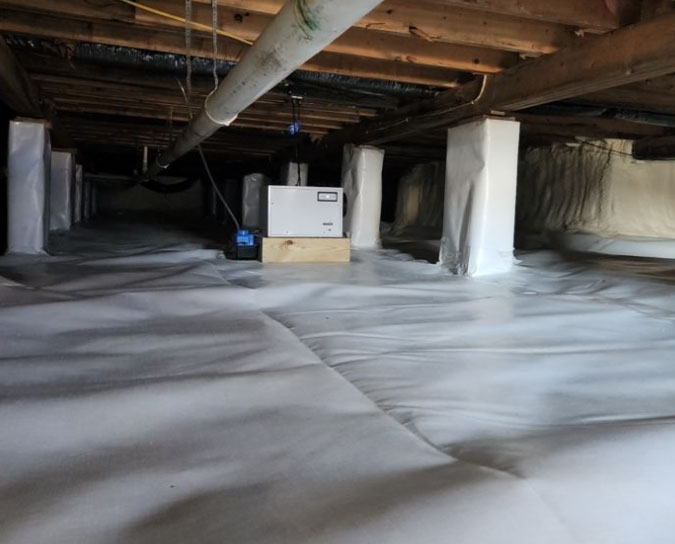 A fully encapsulated crawl space with a thick white vapor barrier across the floor, white-wrapped support piers, spray foam along the rim joists, and an operating commercial dehumidifier.