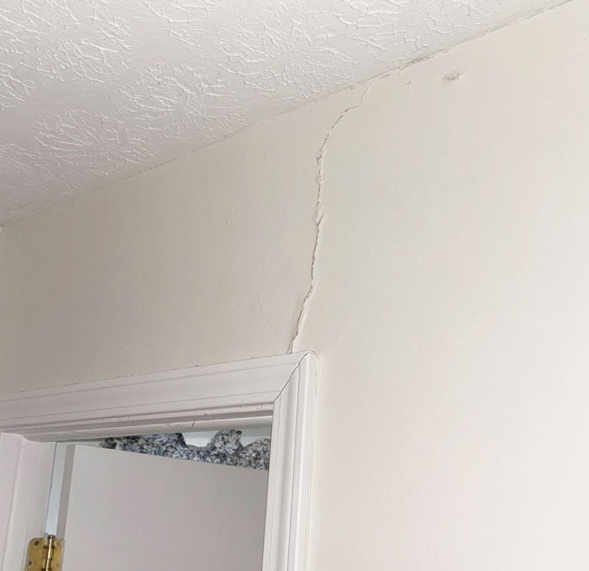 A vertical crack extending upward through interior drywall from the top corner of a door frame to the textured ceiling.