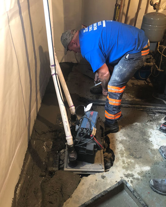 A waterproofing contractor in a blue shirt and work pants smoothing fresh concrete around a newly installed basement sump pump basin and drainage system.