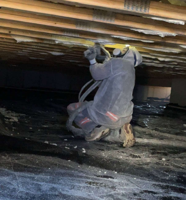 An insulation contractor wearing a full protective suit and mask applying spray foam to subfloor joists while kneeling in a crawl space.