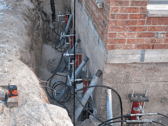 An excavated trench along a brick building foundation showing a series of steel helical piers with hydraulic jacks attached to lift and stabilize the concrete footing.