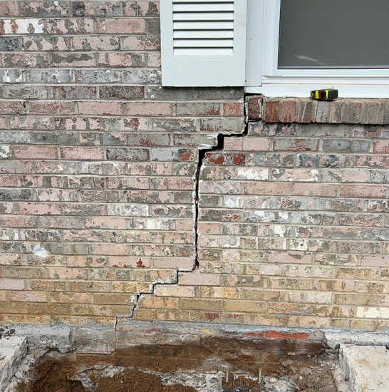 A large stair-step crack in an exterior brick veneer wall beneath a window, with foundation excavation and a metal pier bracket visible at the ground level.