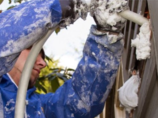 An insulation technician wearing a blue suit injecting white spray foam through a clear hose into an exterior wall opening.
