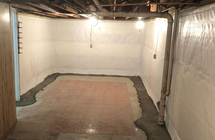 An interior basement room with white vapor barrier panels covering the walls and fresh concrete patch work sealing an interior French drain system along the floor perimeter.