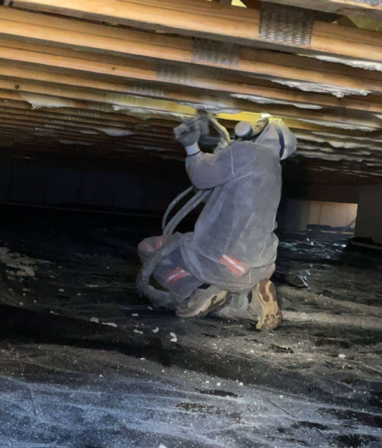 An insulation contractor kneeling on a vapor barrier in a low-clearance crawl space while spraying expanding foam insulation onto wooden subfloor joists.