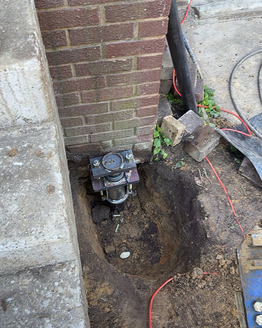 A steel push pier bracket installed in an excavated pit underneath a brick corner foundation next to concrete steps.