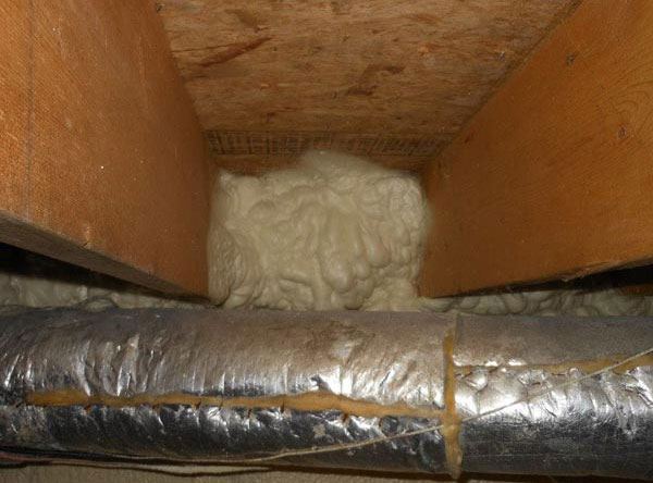 A close-up of spray foam insulation applied to a basement rim joist cavity between wooden floor joists, located behind insulated foil HVAC ductwork.