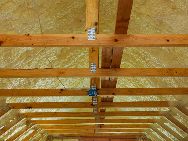 Yellowish-white spray foam insulation applied along the roof deck behind wooden ceiling collar ties and trusses.