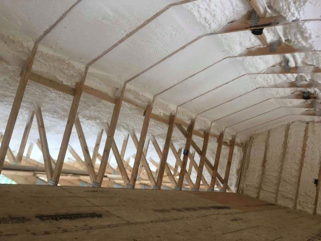 Bright white spray foam insulation applied along the underside of sloped roof rafters and interior end wall studs in a timber-framed attic space.