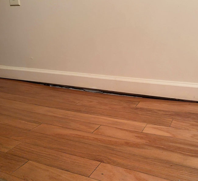 A noticeable gap separating a white baseboard from a sagging wood-look laminate floor along an interior wall.