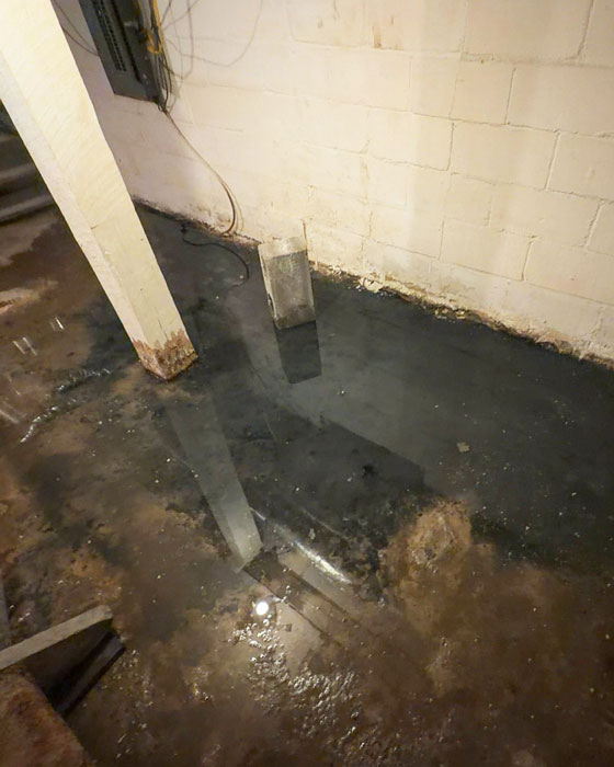 A flooded basement floor with standing dark water reflecting a wooden support post and concrete block foundation wall showing water damage.