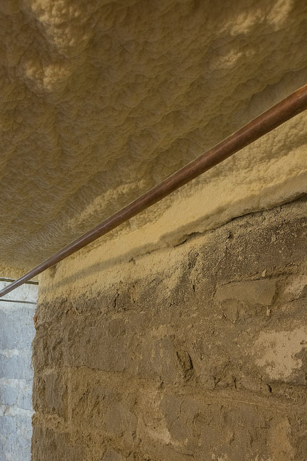 Thick closed-cell spray foam insulation applied to the subfloor ceiling above a rough stone basement foundation wall with a copper pipe running across.