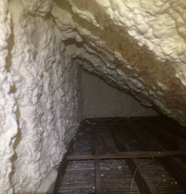 Thick white spray foam insulation lining the sloped ceiling and end gable wall of an attic space above exposed ceiling joists.