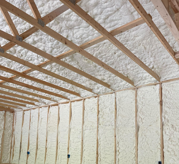 Bright white spray foam insulation thoroughly filling wall stud cavities and sloped roof rafters in a newly framed room.