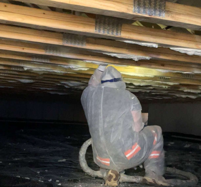 A worker in a full protective suit applies spray foam insulation to wooden floor joists while sitting in a dark crawl space.