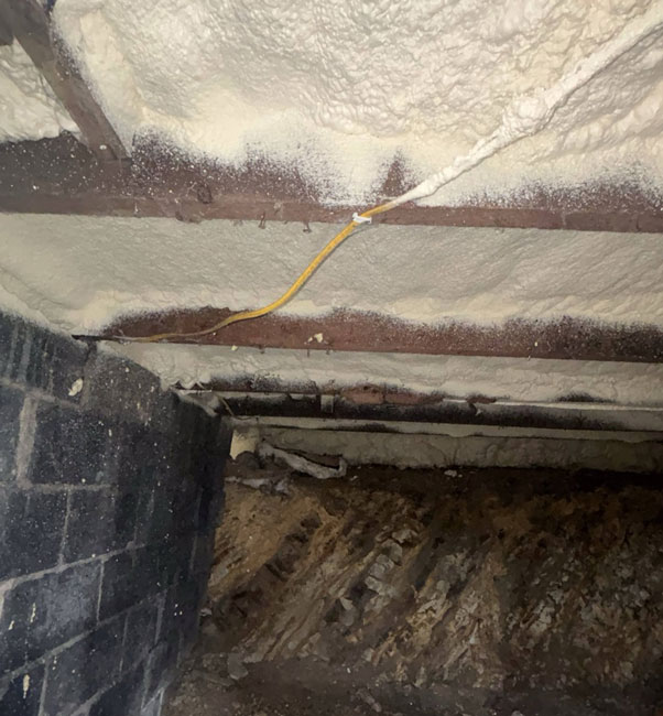 Thick white spray foam insulation applied between dark wooden subfloor joists above a cinder block crawl space wall and exposed earth floor.
