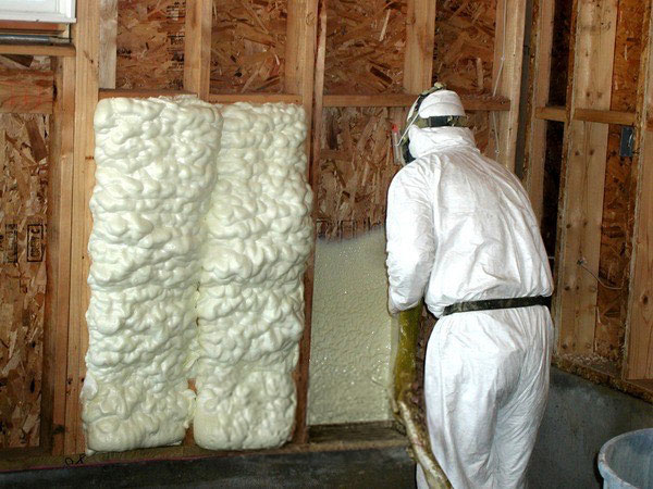 An insulation technician wearing a white protective suit and mask sprays expanding foam into a wooden wall stud cavity.