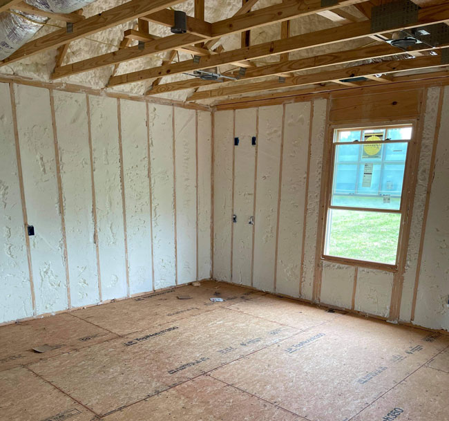 Bright white spray foam insulation filled between wall studs in a framed interior room with OSB subflooring and open roof trusses.