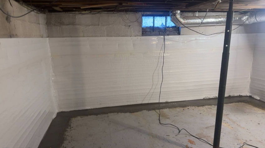 A basement corner with bright white vapor barrier paneling lining concrete block walls above a newly cemented floor drainage perimeter, featuring a support post and ductwork.