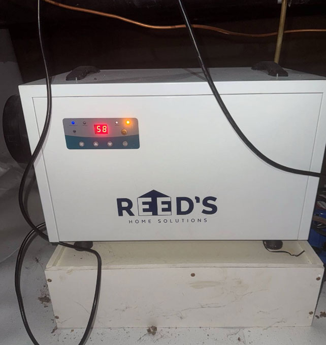 A white Reed's Home Solutions commercial dehumidifier unit with a digital display reading 58, set on an elevated box inside an encapsulated crawl space.