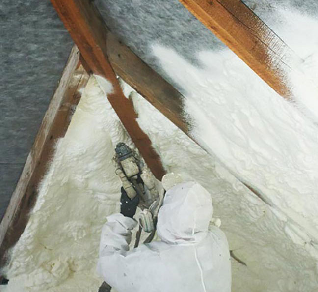 An insulation technician wearing a protective white suit, hood, and respirator sprays white foam onto sloped roof decking between wooden rafters.